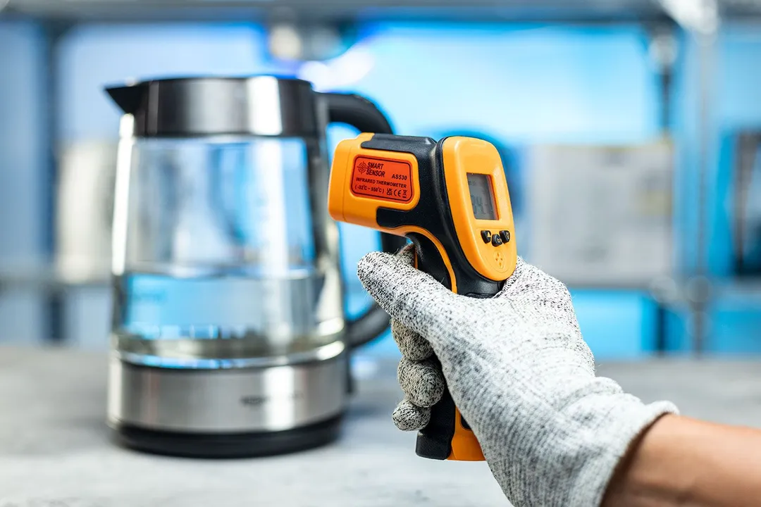 The Smart Sensor AS530 Infrared Thermometer in the gloved hand of a reviewer against a blurry blue backdrop with a kettle.