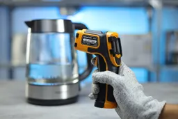 The SOVARCATE HS960D Infrared Thermometer in the gloved hand of a reviewer against a blurry blue backdrop with a pitcher.