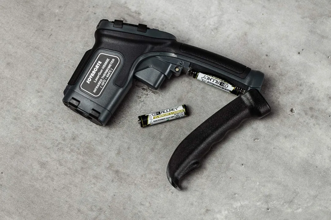 The Sovarcate HS980E IR thermometer with its battery compartment opened and one of the AAA batteries slotted in. A spare battery is to the left.