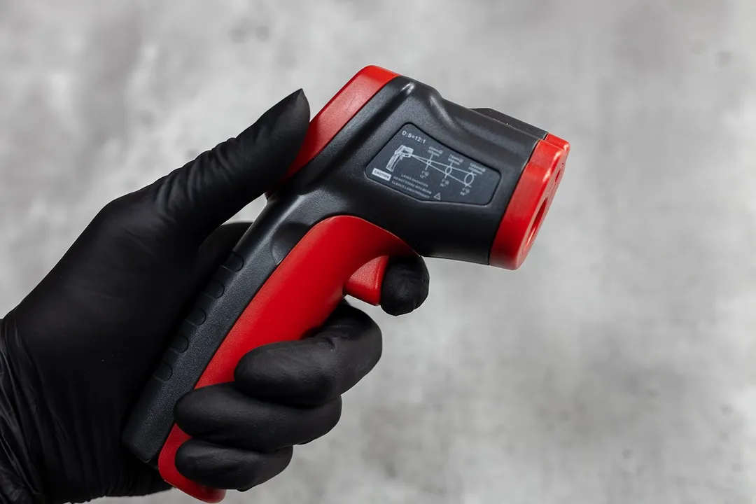 The Wintact WT530 IR thermometer in the black-gloved hand of a reviewer and a finger on the red trigger of the IR thermometer.