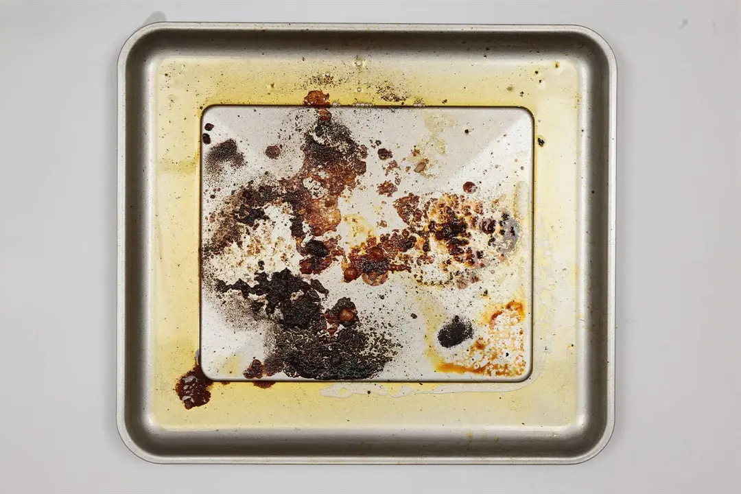 Caramelized meat juice and grease on the silver baking pan of the Cuisinart TOA-60 Convection Toaster Oven Air Fryer on a grey background.