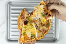 Two hands break apart a slice of baked pizza using a toaster oven with melty cheese, meat, and green bell peppers on top. In the background is a pizza on top of a grooved baking tray.