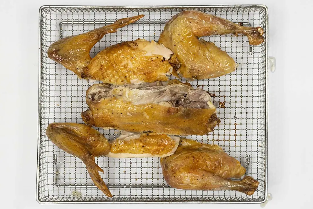 The golden brown roasted whole chicken is carved into seven pieces inside an air fryer basket with a silver baking pan below. There’s a carcass, two breast pieces, two wings, and two thighs.