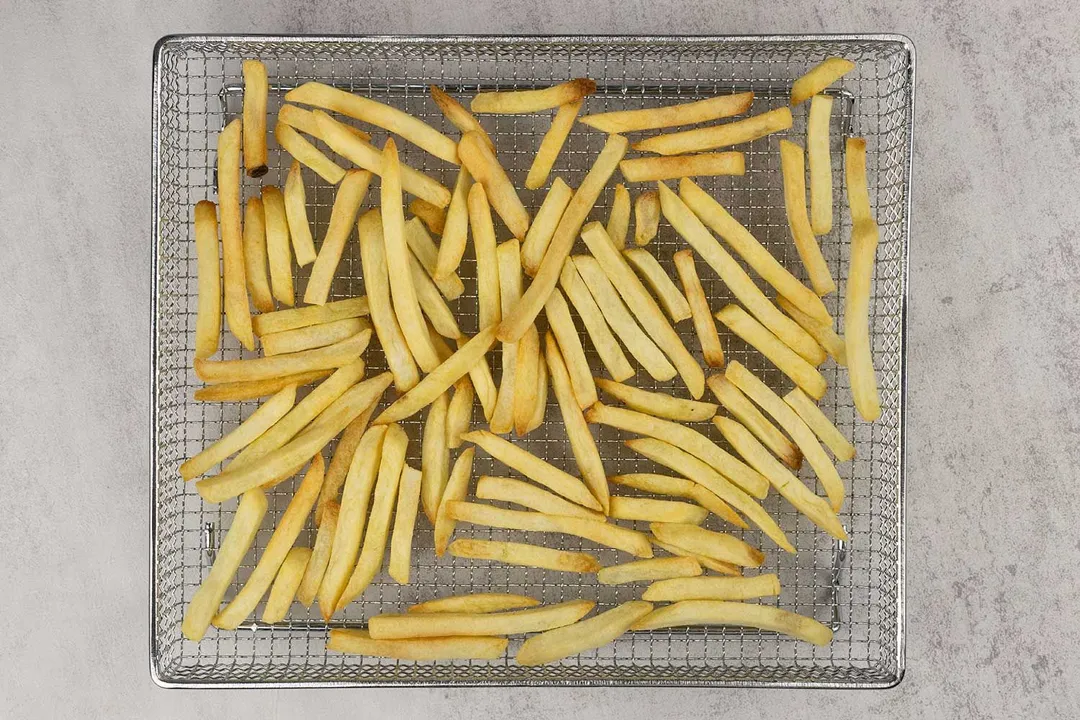 Pieces of golden baked french fries using a toaster oven inside a stainless steel air fryer basket on a grey background.