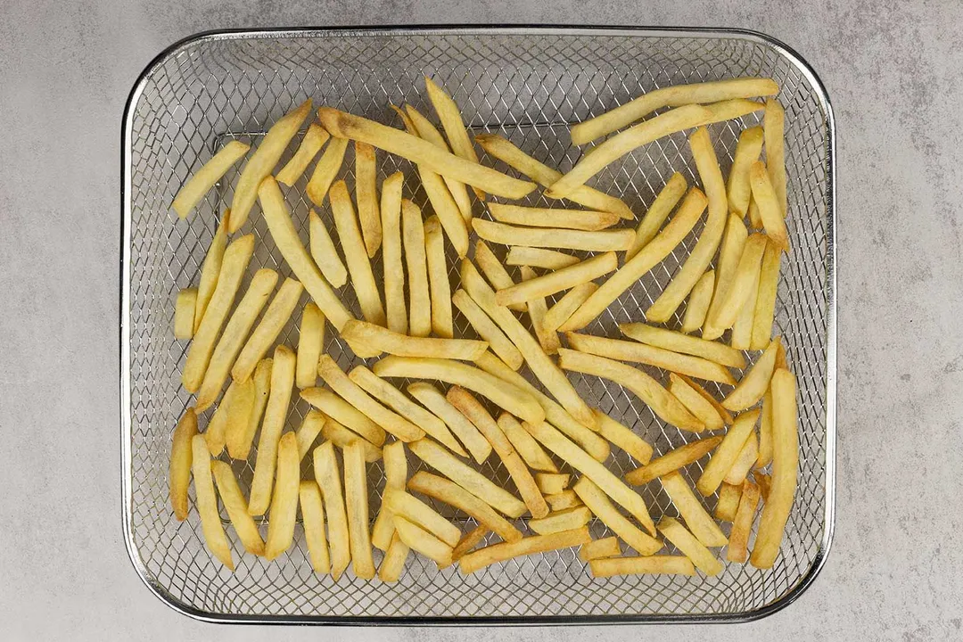 Pieces of baked french fries using a toaster oven inside an air fryer basket on a grey background.