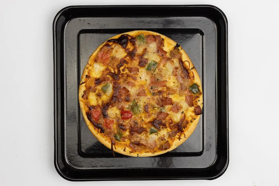 A toaster oven baked 9-inch thick-crust meat pizza with cheese, onions, and green bell peppers on top inside a baking pan.