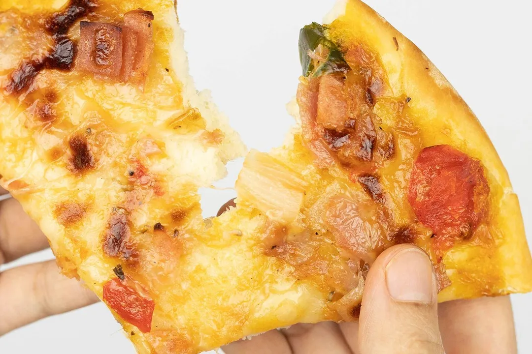 Two hands break apart a slice of pizza with melty cheese, meat, and green bell peppers on top baked using a toaster oven.