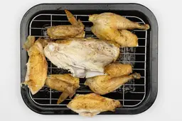 Seven pieces of carved chicken including a carcass, two breasts, two wings, and two thighs on a broiling rack and baking pan.