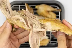 Two hands pulling apart a roasted chicken thigh. In the white background is the rest of the roasted chicken on a baking pan.