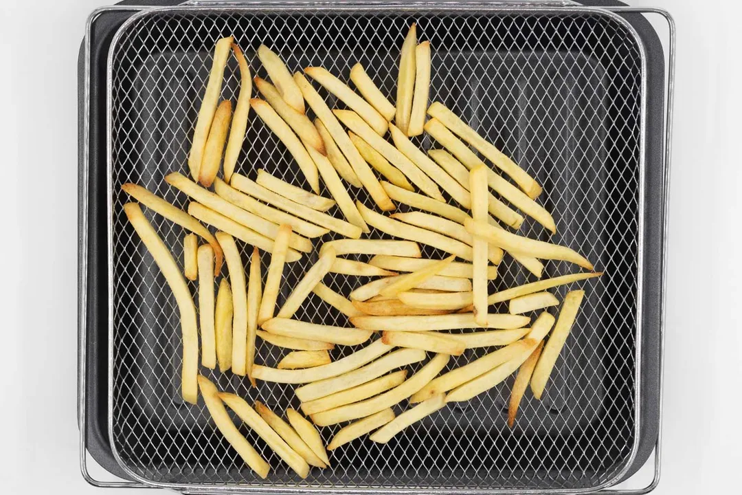 Pieces of baked french fries using the Cosori CO130-AO inside an air fryer basket on a grey baking pan on a white background.