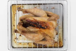 A whole roasted chicken using the Hamilton Beach 31344DA belly up on an oven rack and baking pan on a white background.
