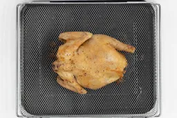 A whole roasted chicken using the Ninja DT201 backside up inside an air fryer basket and baking pan on a white background.
