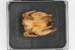 A whole roasted chicken using the Ninja DT201 belly up inside an air fryer basket and baking pan on a white background.