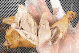 A hand holding a pulled-apart roasted chicken thigh. In the background are an air fryer basket and baking pan.