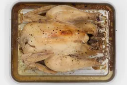 A whole roasted chicken using the Mueller MT-175 Toaster Oven belly up on a silver baking pan on a white background.