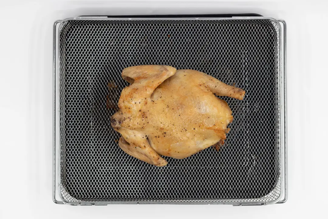 A whole roasted chicken using a toaster oven on a white background. The chicken is backside up inside an air fryer basket on top of a black baking pan.