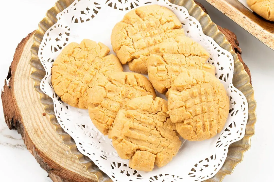 Six golden cookies on top of a paper lining in a serrated dish on top of a wooden tray resembling the cross surface of a log.