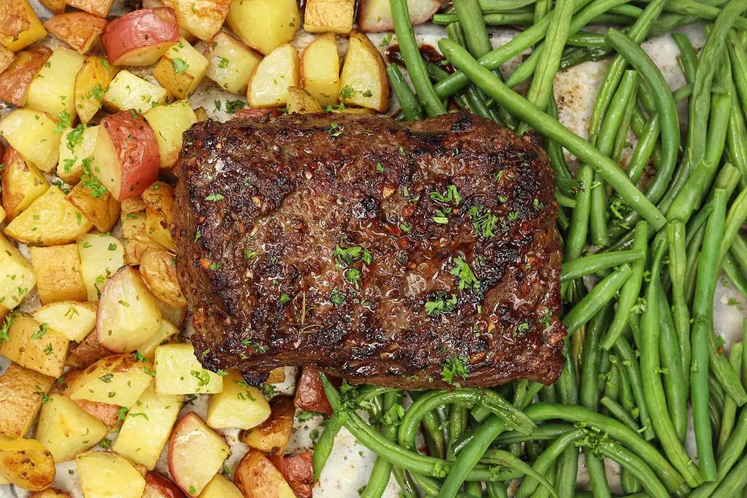 In the middle is a piece of seared steak with parsley on top, beneath the steak on the left are baked pieces of potato and on the right are baked green beans.