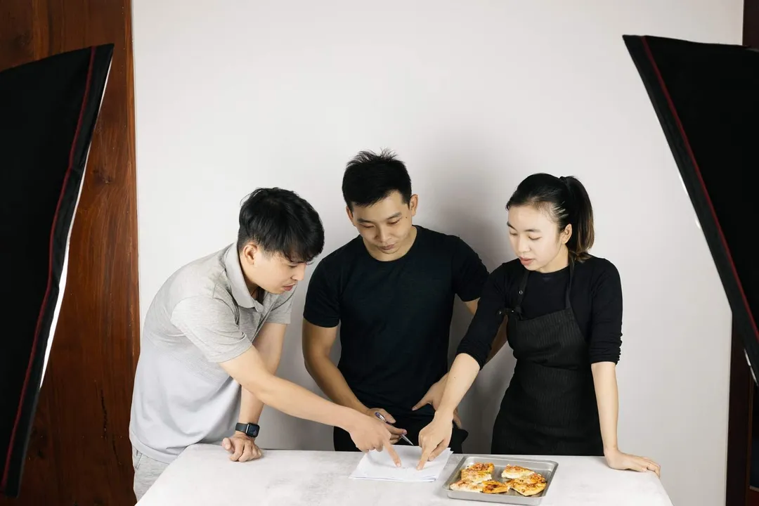 Our team of three people is writing the scores for a baked pizza using a toaster oven on a sheet of paper using a pen. The background is grey, with two lights on the sides.
