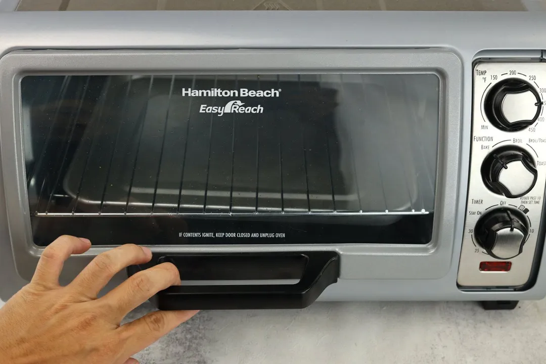 A front view of a hand holding the roll top door handle of the Hamilton Beach 31127D 6-Slice Easy Reach Toaster Oven.