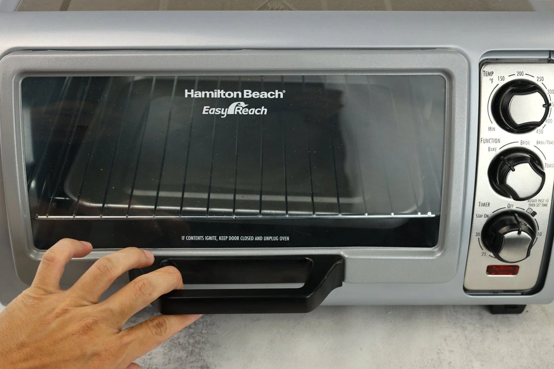 A front view of a hand holding the roll top door handle of the Hamilton Beach 31127D 6-Slice Easy Reach Toaster Oven.