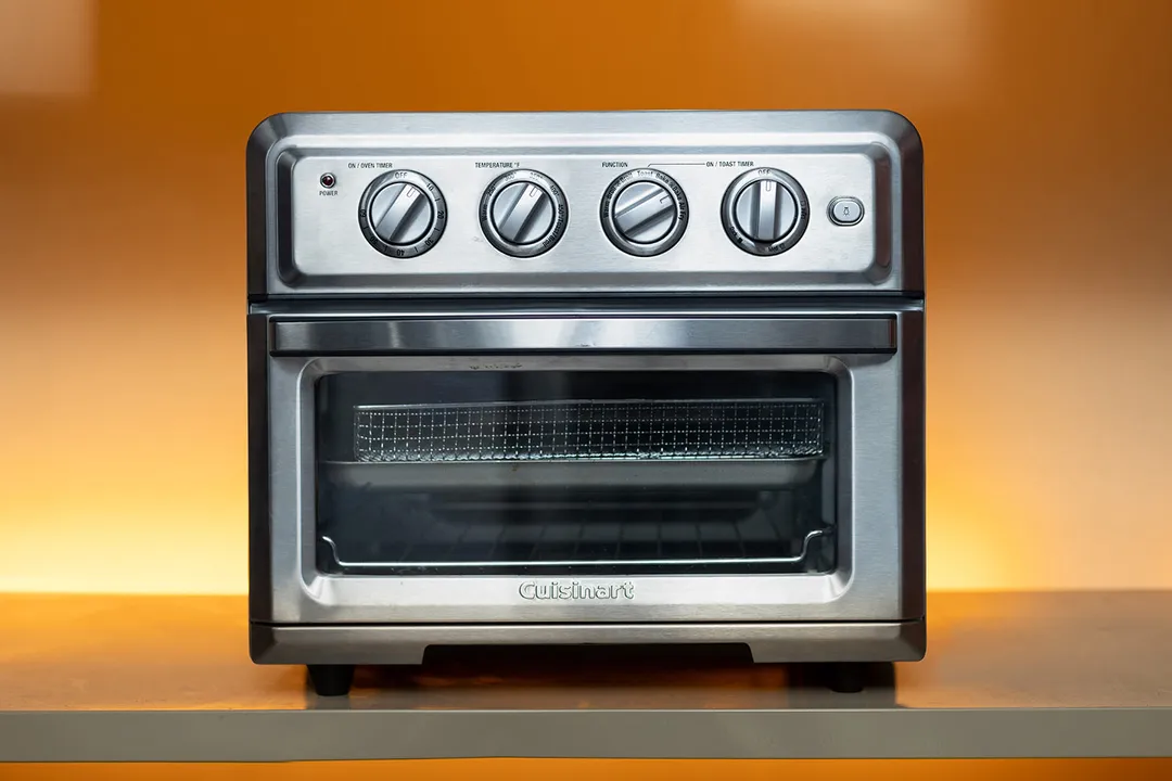 The front of a closed Cuisinart TOA-60 Convection Toaster Oven Air Fryer. The control panel is positioned horizontally on the upper front. Below the control panel is the door handle. The glass door is covered with an illustration.
