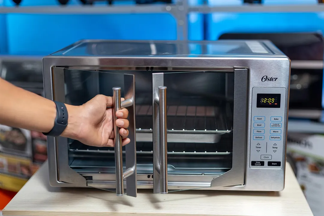 The front of a closed Oster TSSTTVFDDG XL Digital French Door Convection Toaster Oven. The control panel is positioned vertically on the right. Inside the oven are a shining light, two baking racks, and one baking pan.