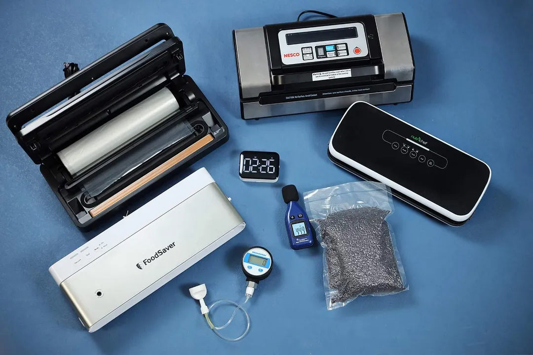 A collection of best vacuum sealers and testing instruments on a blue surface. There’s a vacuum gauge, a sound level meter, a timer, and a vacuum sealed bag with black rice grains.