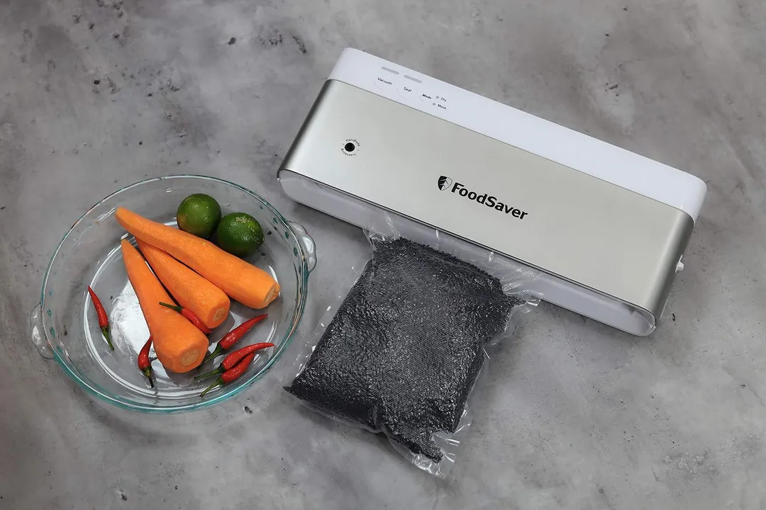 The FoodSaver VS-0160 sits next to a glass bowl containing carrots, limes, and red chili peppers. In front of the sealer is a packed plastic bag of black rice grains.