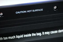 The “Caution: Hot Surface” warning sticker located near the heat bar within the interior of the Mueller MV-1100 sealer.