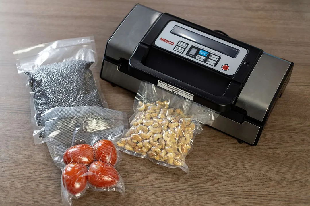 Nesco VS-12 Deluxe vacuum sealer packing a bag of roasted cashews. In front of it are packed bags of tomatoes and black rice grains.