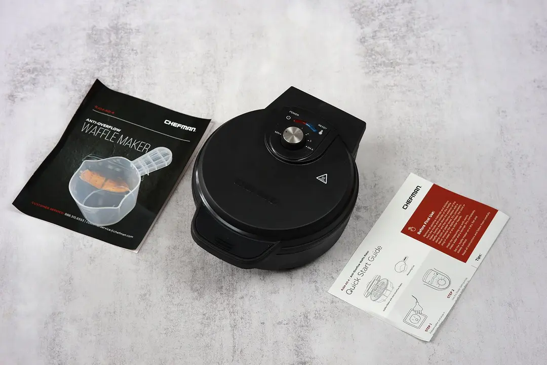 The all-black Chefman waffle maker next to its instruction manual and accessory (a cup for measuring batter).
