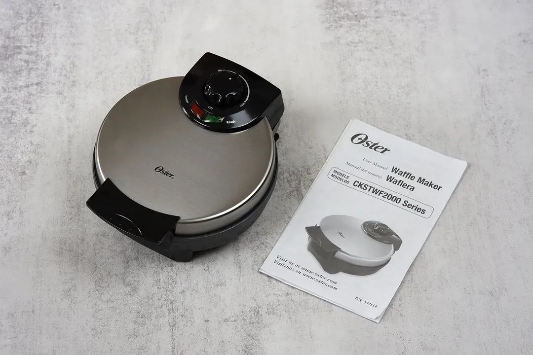The Oster waffle maker, with its stainless steel top lid and black plastic detailing, is placed next to its manual.
