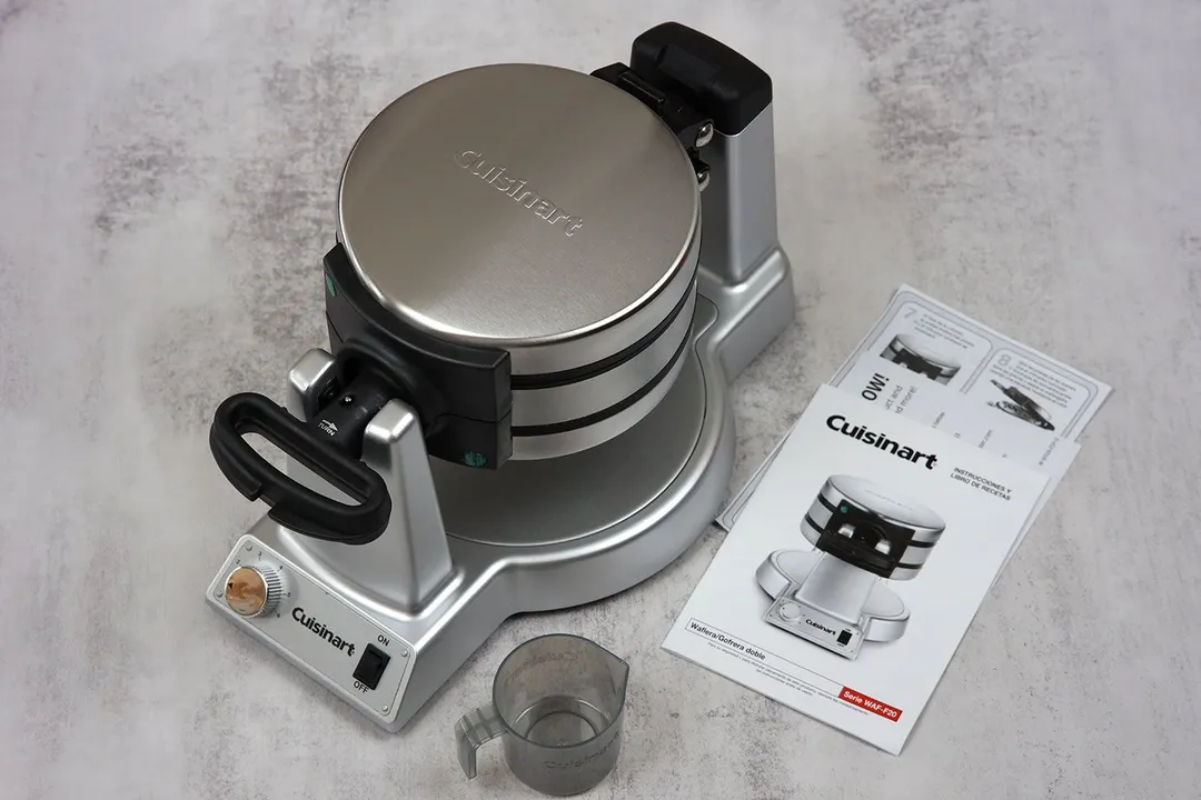 The silver Cuisinart WAF-F20P1 Double waffle maker sitting on a table next to its user manuals and a measuring cup.