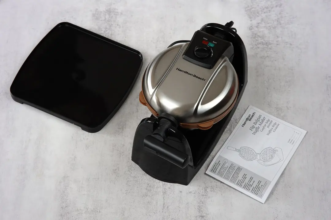 The Hamilton Beach Flip Belgian waffle maker (26031) is on a table next to the included drip tray and the user manual.