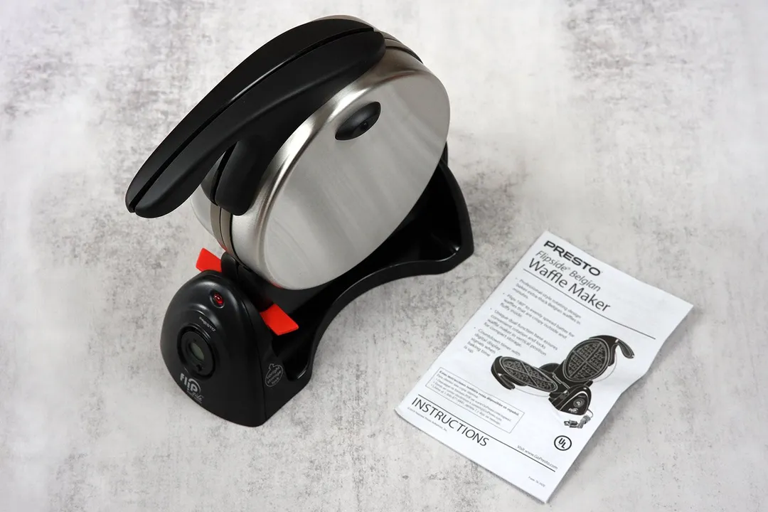 The Presto Belgian waffle maker with black-and-red plastic detailing and glossy metallic body next to its instruction manual.