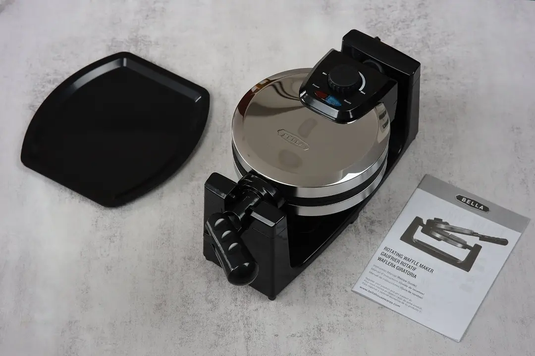 The Bella stainless steel waffle maker (13991) sitting next to its instructional manual and the black removable drip tray.