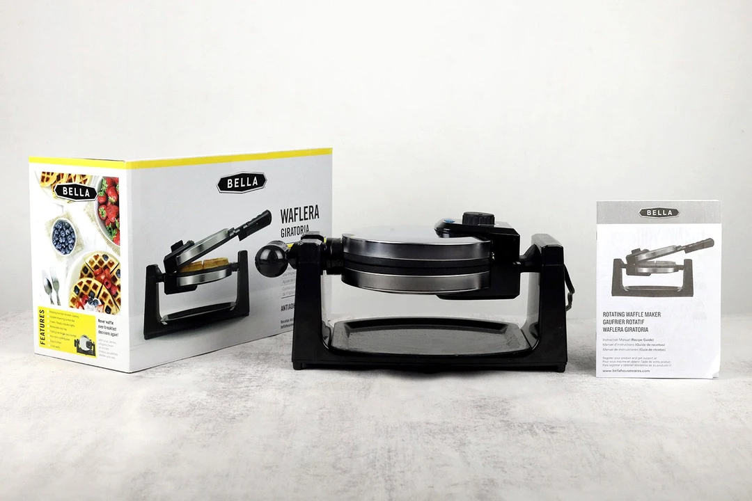 The BELLA rotating waffle maker with its glossy, metallic baking chamber positioned next to its instruction manual and shipping box.