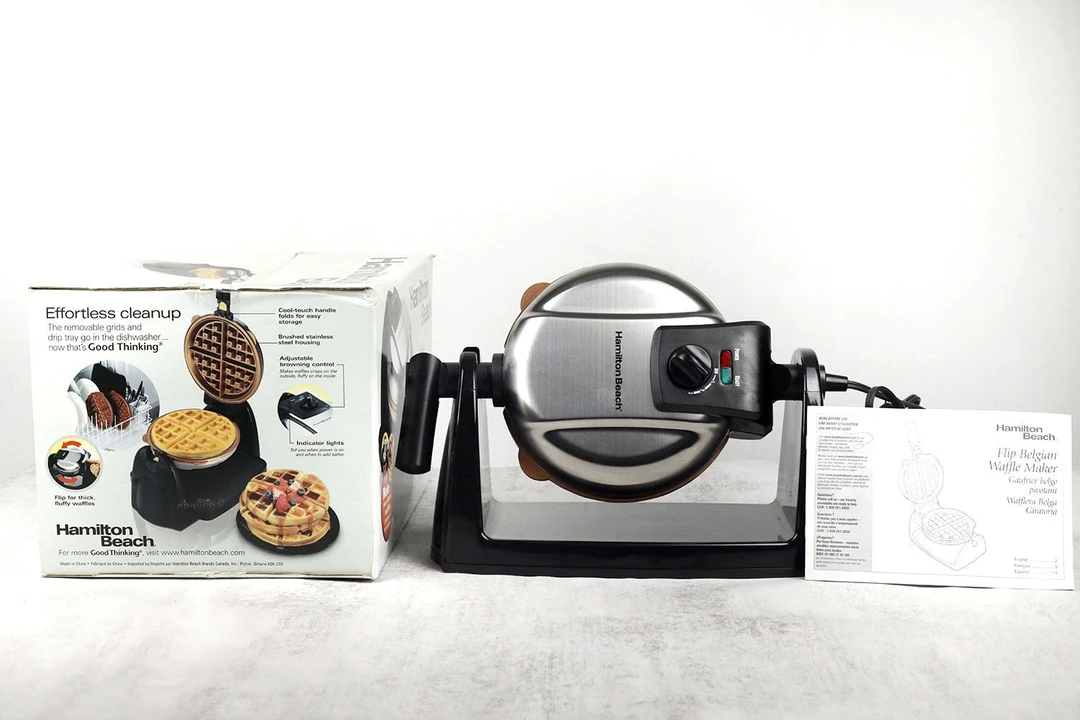 The silver Hamilton Beach 26031 Belgian waffle maker presented on a table alongside the user manual and its shipping box.