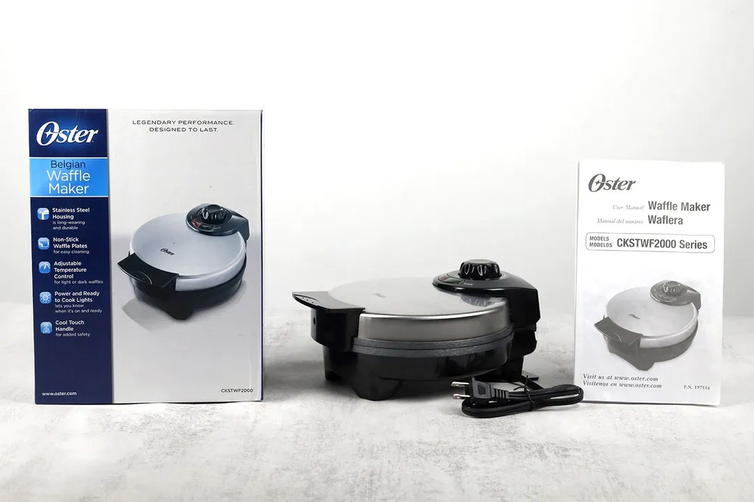 The Oster waffle maker, with its metal lid and black plastic base, sits amidst its shipping box (left) and manual (right).