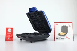 The Dash No-Drip waffle maker with its lid opened, revealing its black waffle plates. To either side are instruction manuals.