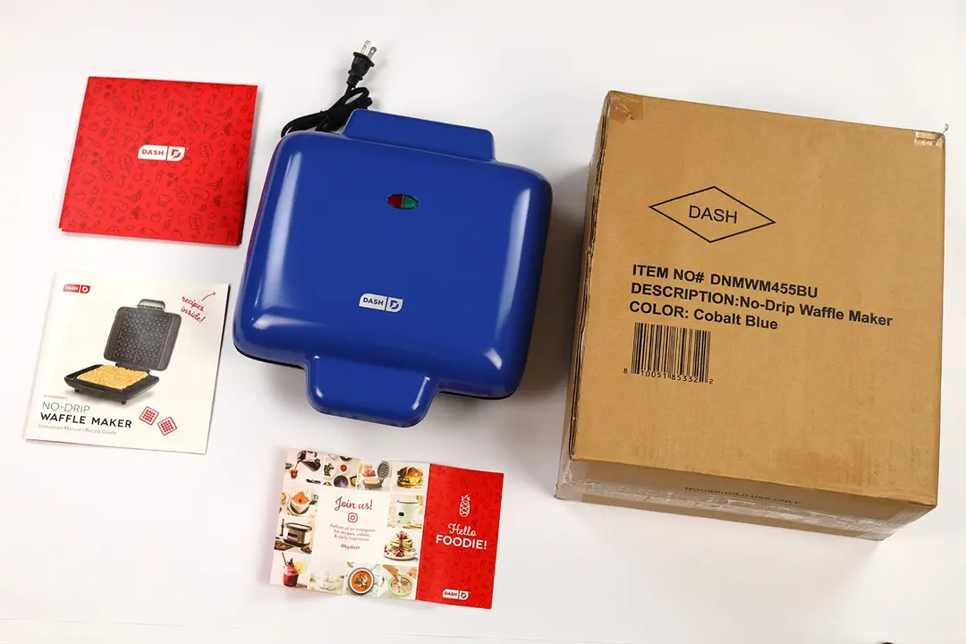 The blue Dash No-Drip waffle maker next to its shipping box and instruction manuals.