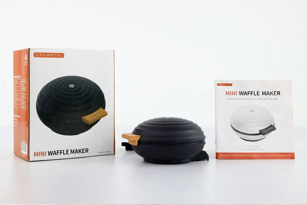 The black CROWNFUl waffle maker with wood-tone handle on a table next to its instructional manual and the shipping box.
