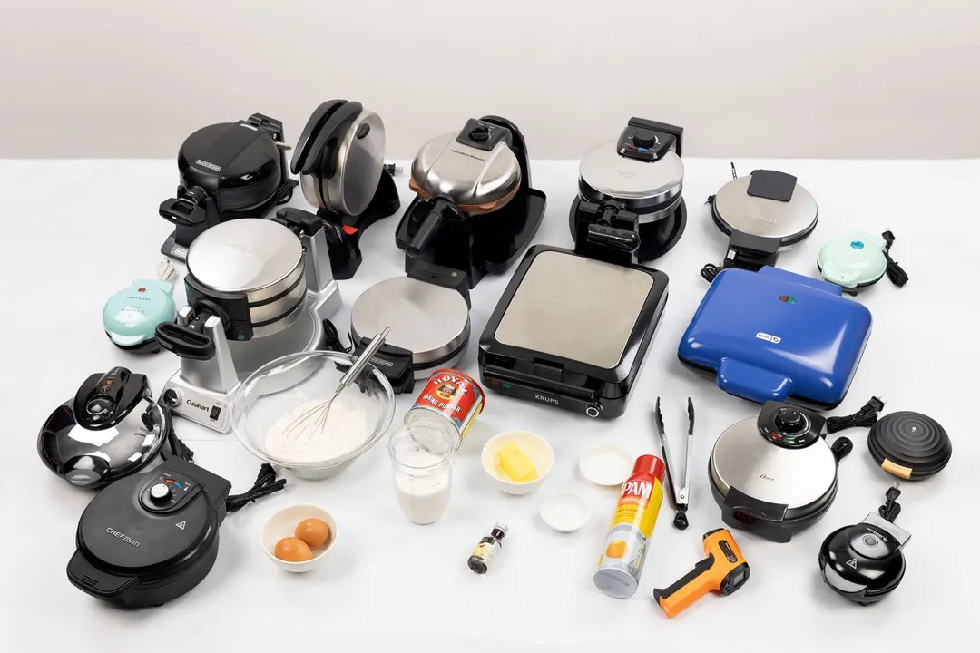 Waffle makers arranged on a table next to bowls containing flours, eggs, butter, and other ingredients for mixing waffles. Cooking tools like tongs and testing equipment such as thermometers.