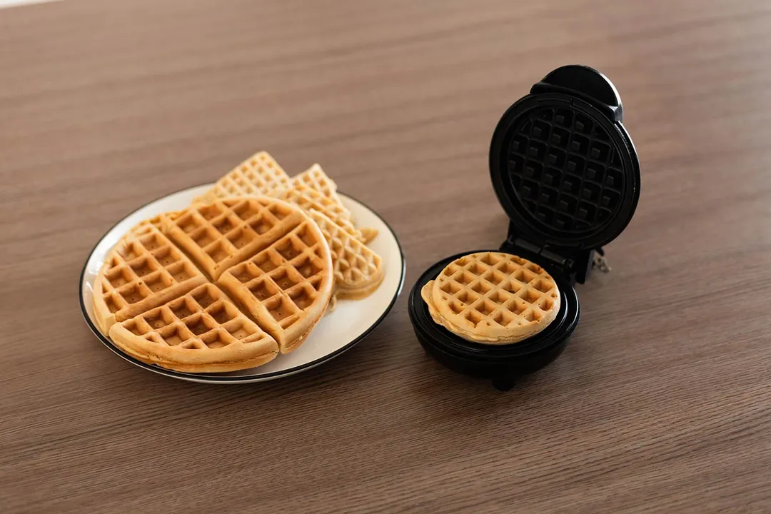 The Holstein Housewares Mini waffle maker next to a plate of cooked mini and 4-inch waffles on a wooden tabletop.