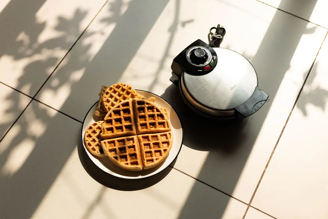 The Oster waffle maker on a wooden floor next to a plate of 7-inch and mini waffle makers.