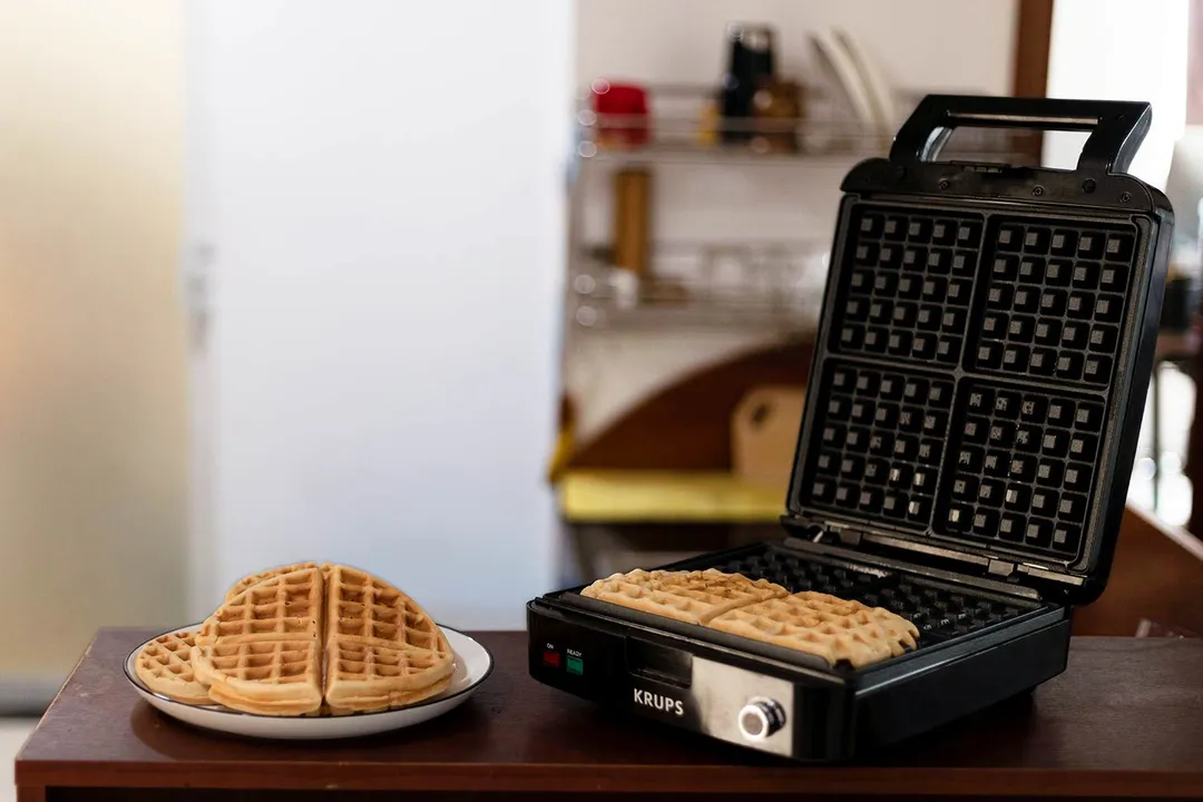 The KRUPS Belgian waffle maker with its lid opened and two waffles on the plate next to a plate of cooked waffles on a wooden countertop