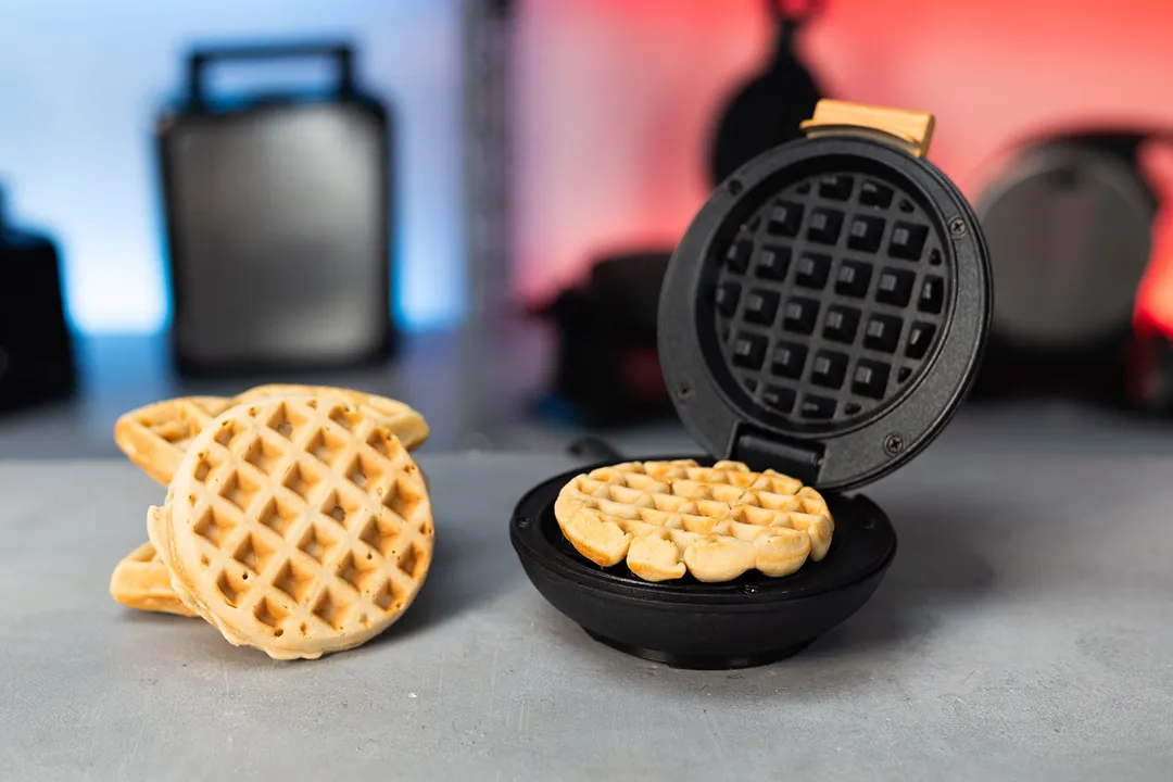 Crownful Mini waffle maker with its lid opened and a fresh waffle inside. Next to it is a stack of mini waffles.