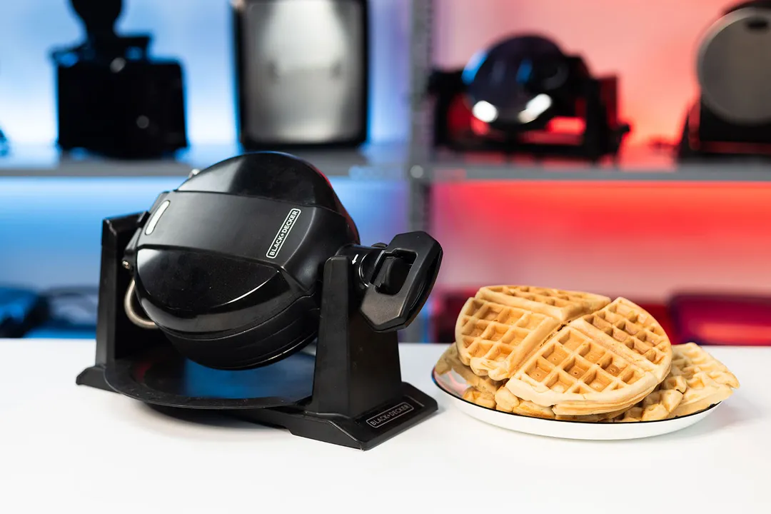The Black and Decker Double waffle maker with a plate of fresh waffles beside it on a white countertop against a blurry red and blue background.