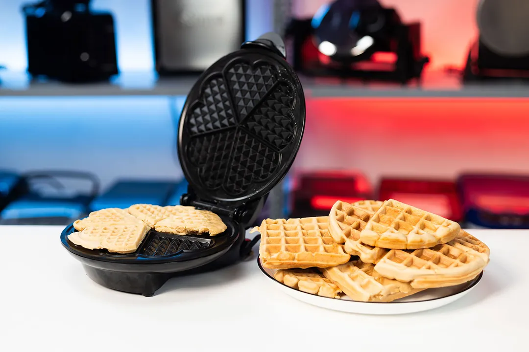 Euro Cuisine Heart-Shaped waffle maker with opened lid revealing its unique heart-shaped plates on a white countertop next to a plate of fresh waffles.